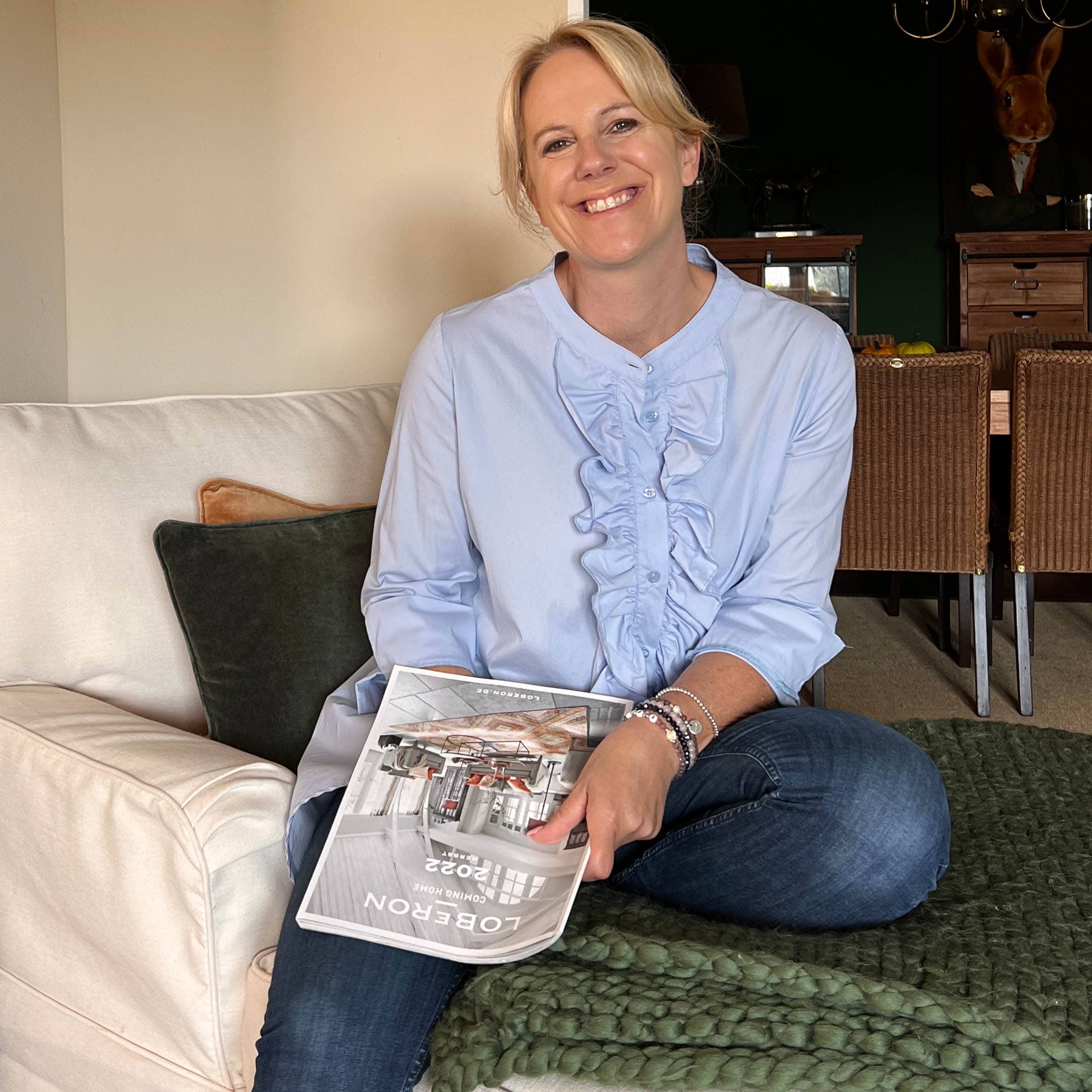 Frau sitzt auf dem Sofa mit einem Loberon-Magazin, gemütliches Zuhause mit grüner Strickdecke und Kissen.