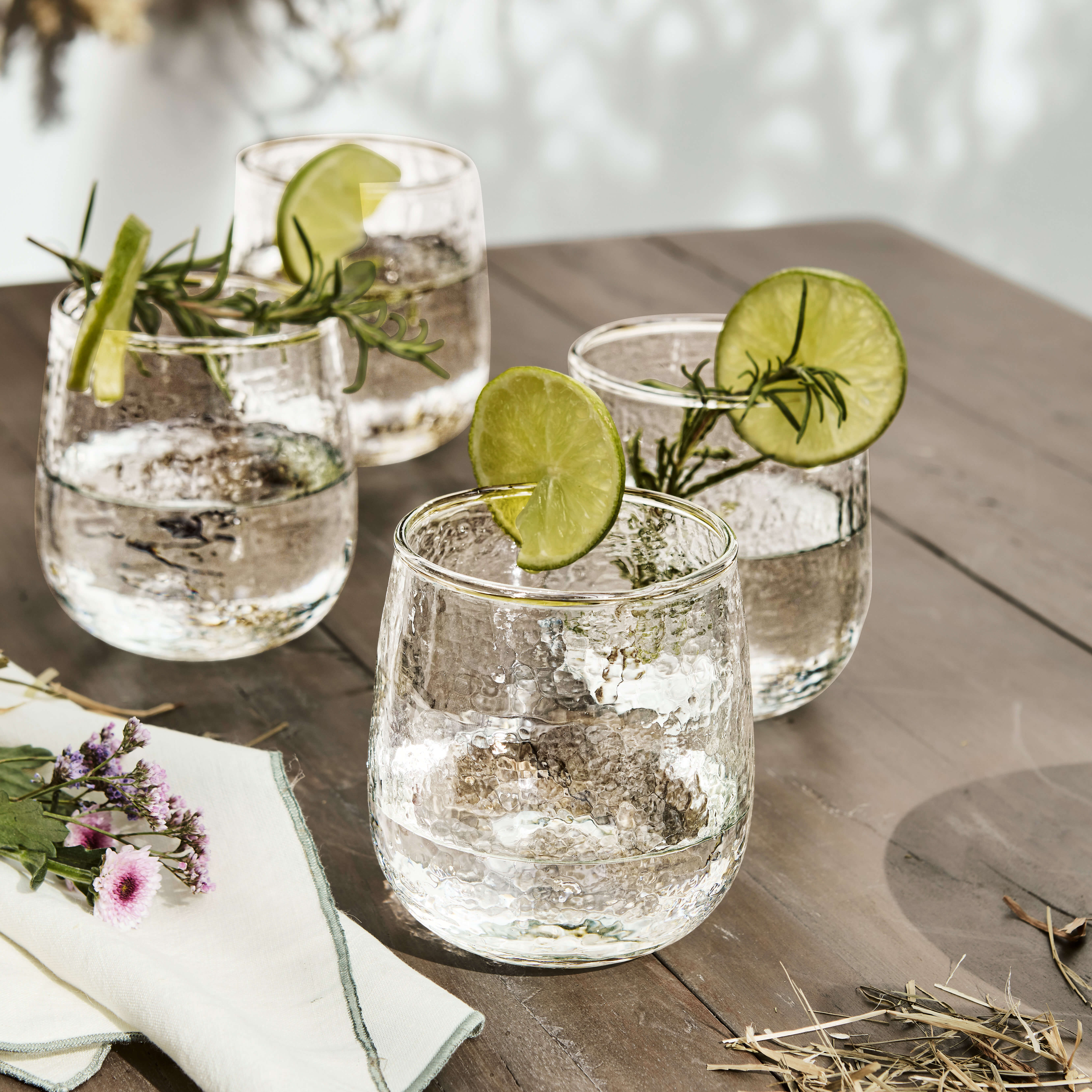 Vier strukturierte Gläser mit Wasser, Limettenscheiben und Rosmarinzweigen auf einem Holztisch arrangiert