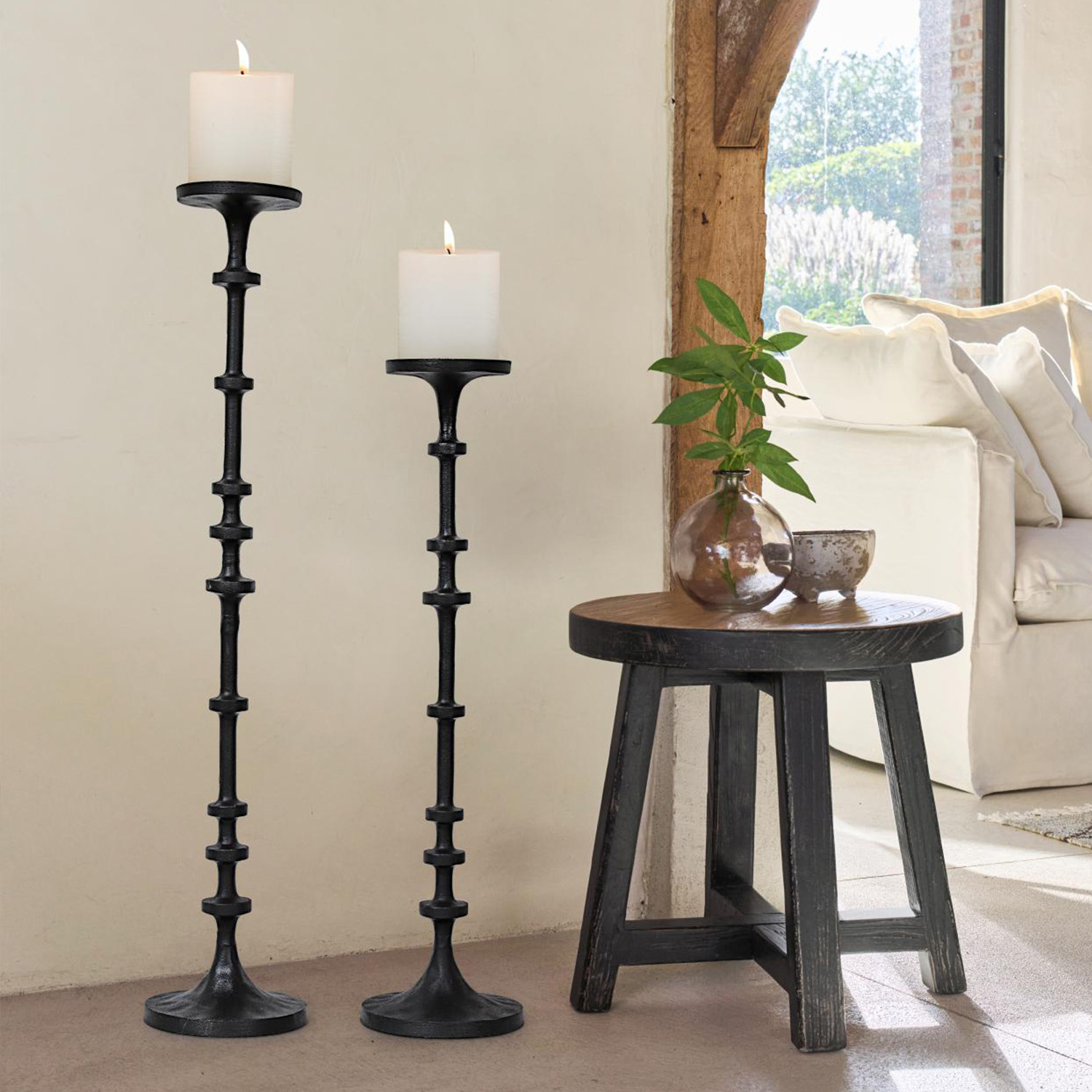 Zwei schwarze Metall-Kerzenhalter mit weissen Kerzen neben einem Holzhocker und Sofa im Wohnzimmer
