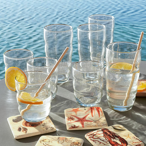 Gläser aus klarem Glas mit Zitronenscheiben und Holzrührstäbchen, maritime Untersetzer am Wasserufer