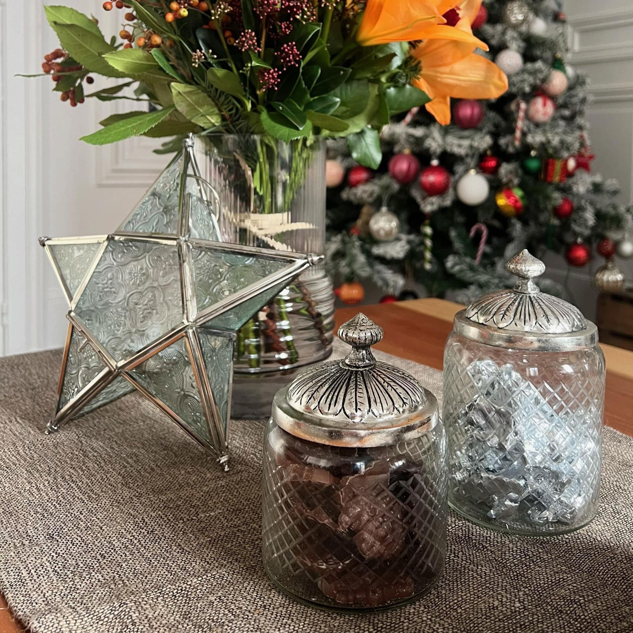 Gläser mit Metalldeckeln und dekorativem Stern auf dem Tisch mit Blumen und Weihnachtsbaum im Hintergrund