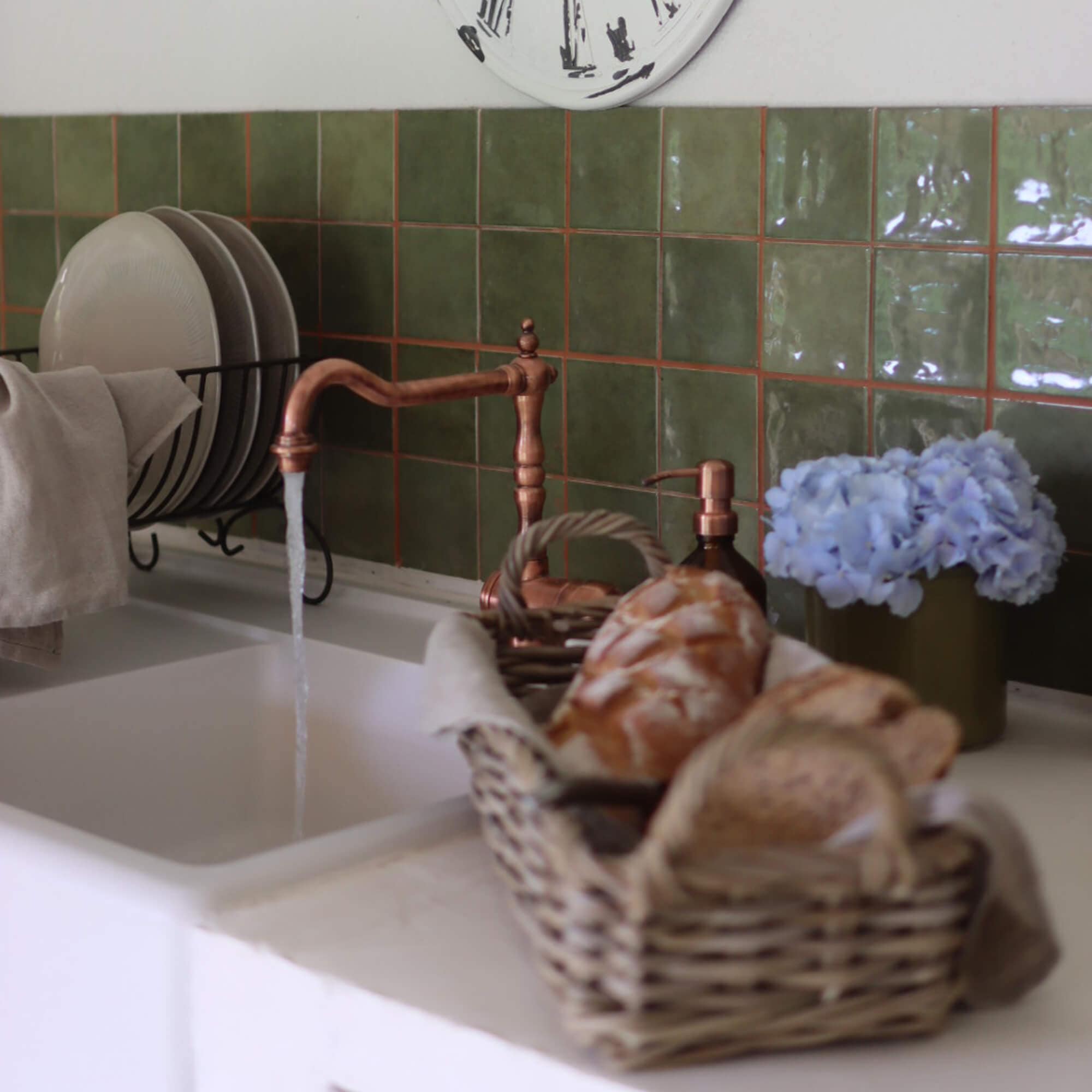 Küche mit kupferfarbenem Wasserhahn, grünen Fliesen, Brotkorb und blauen Blumen als Dekoration