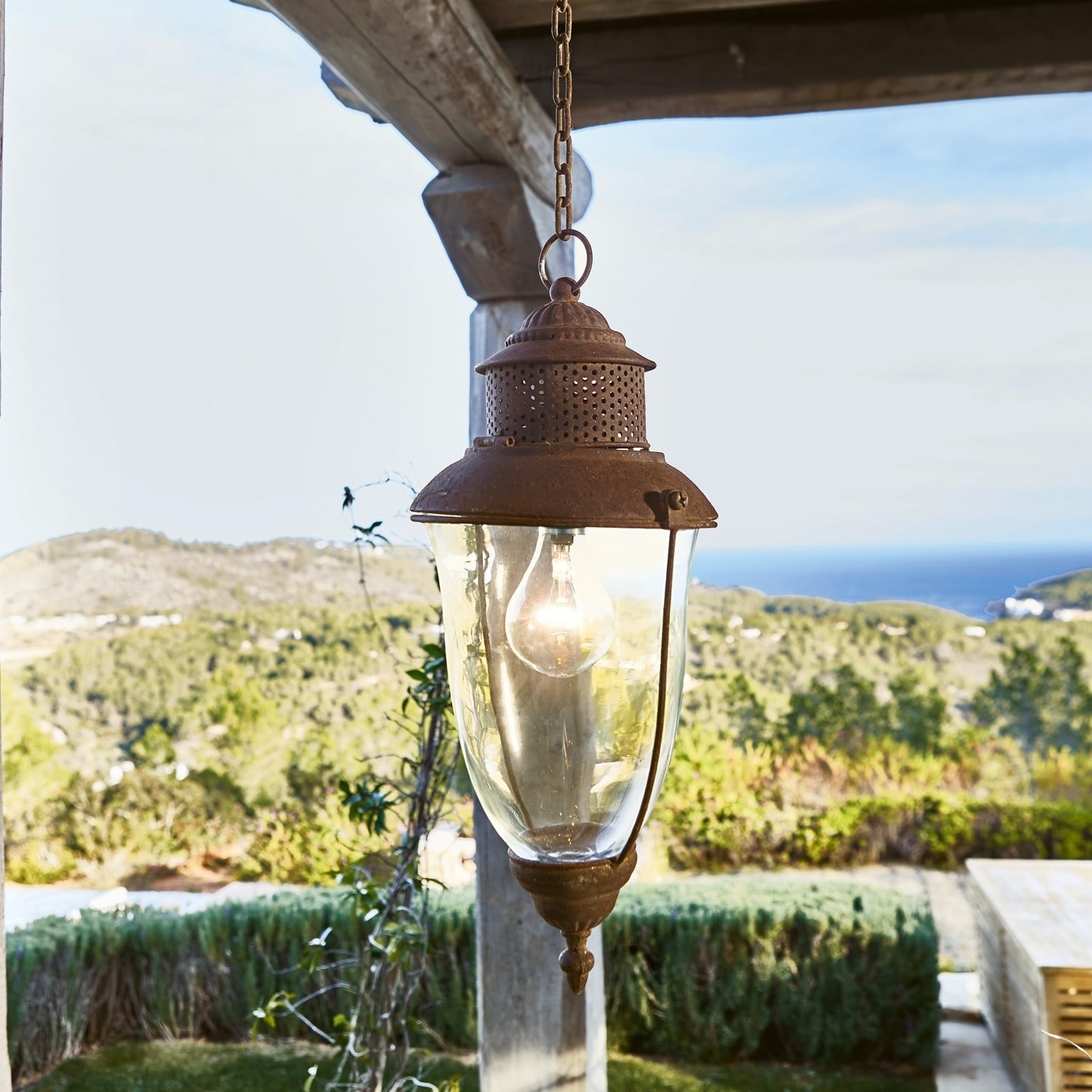 Hängelaterne Floricia Hängende Laterne aus rostigem Metall und Glas mit Blick auf grüne Hügel und das Meer im Hintergrund