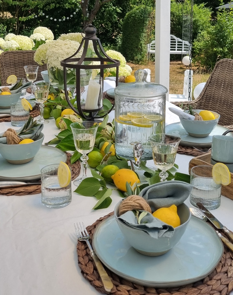Sommerlicher Gartentisch mit blauer Keramik, Zitronen und Glaswasserbehälter mit Zitrone