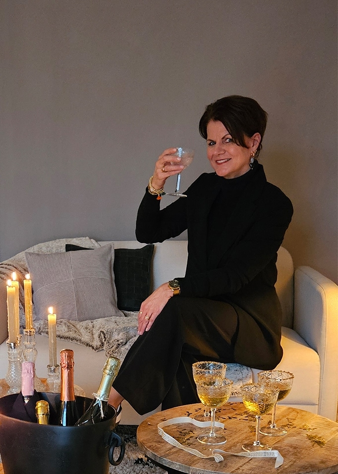 Frau in schwarzer Kleidung sitzt auf hellem Sofa, daneben Glas, Kerzen und Champagnerflaschen.