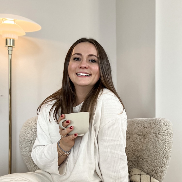Lächelnde Frau im weissen Pullover hält eine beige Tasse, sitzt auf weichem Sessel in hellem Raum
