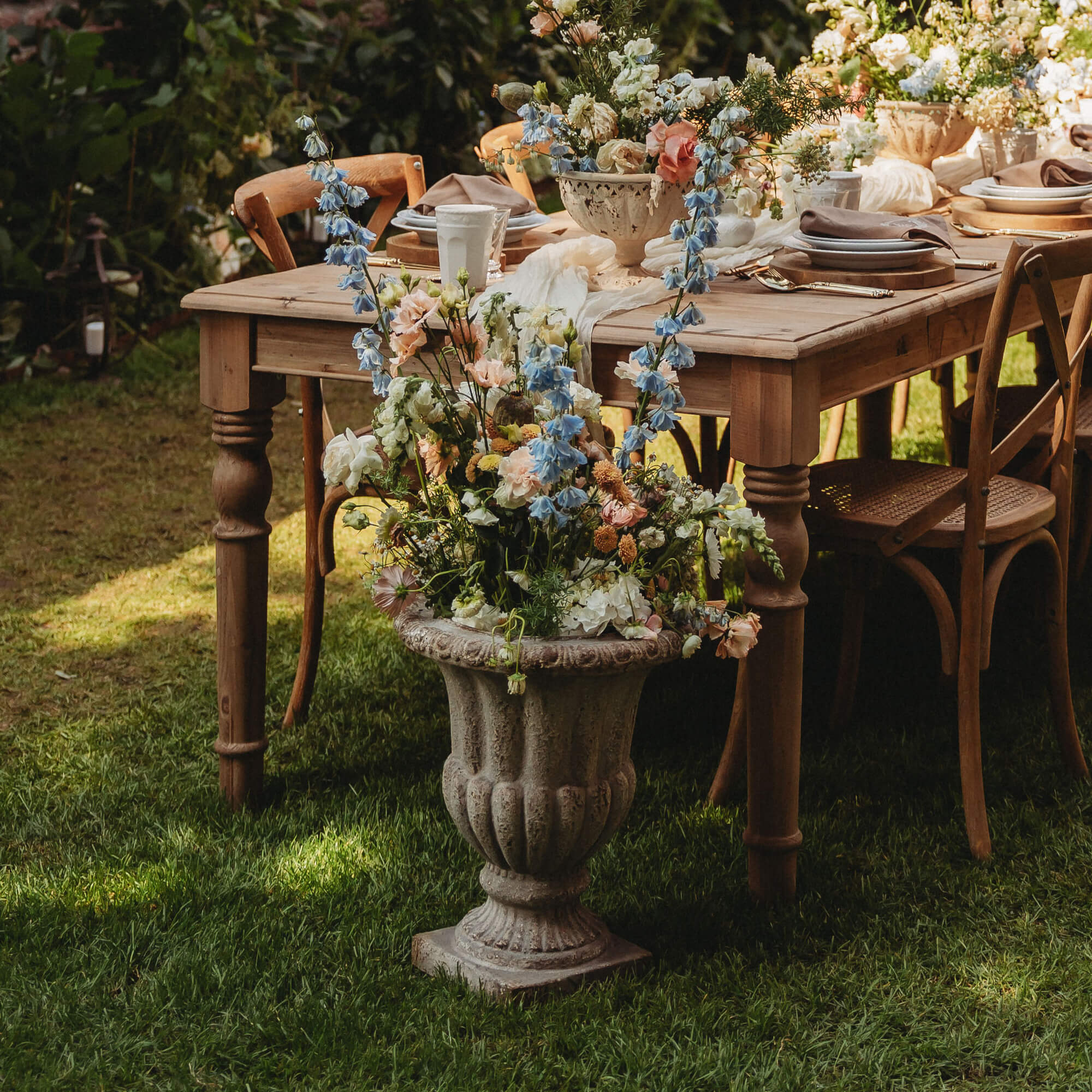 Grosse steinerne Blumenvase mit pastellfarbenen Blumen vor einem Holztisch und Stühlen im Garten
