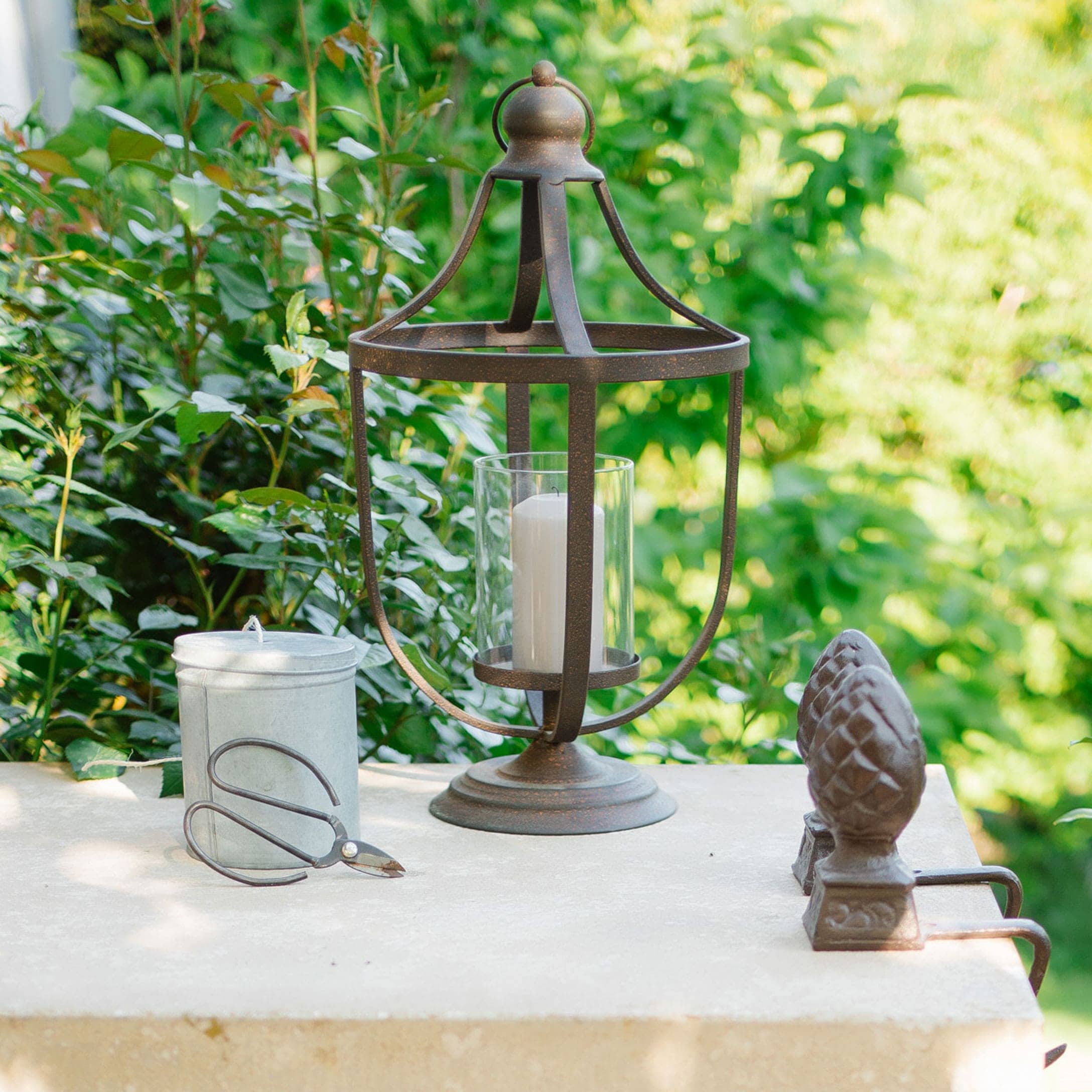 Metalllaterne mit weisser Kerze und Gartenschere auf hellem Tisch vor grünem Hintergrund