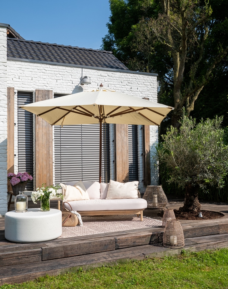 Terrasse mit weisser Ziegelwand, beiges Sofa mit Kissen und cremefarbenem Sonnenschirm auf Holzdeck