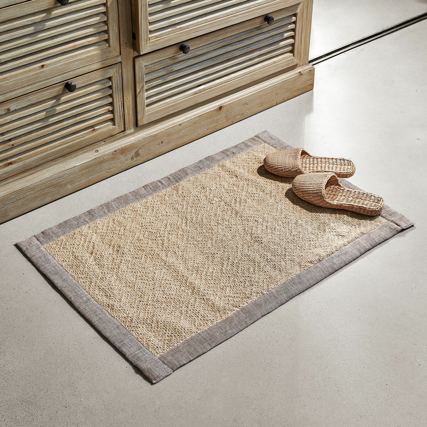 Badematte Wilder Beiger Teppich aus Naturmaterial mit grauem Rand auf hellem Boden neben Holzkommode
