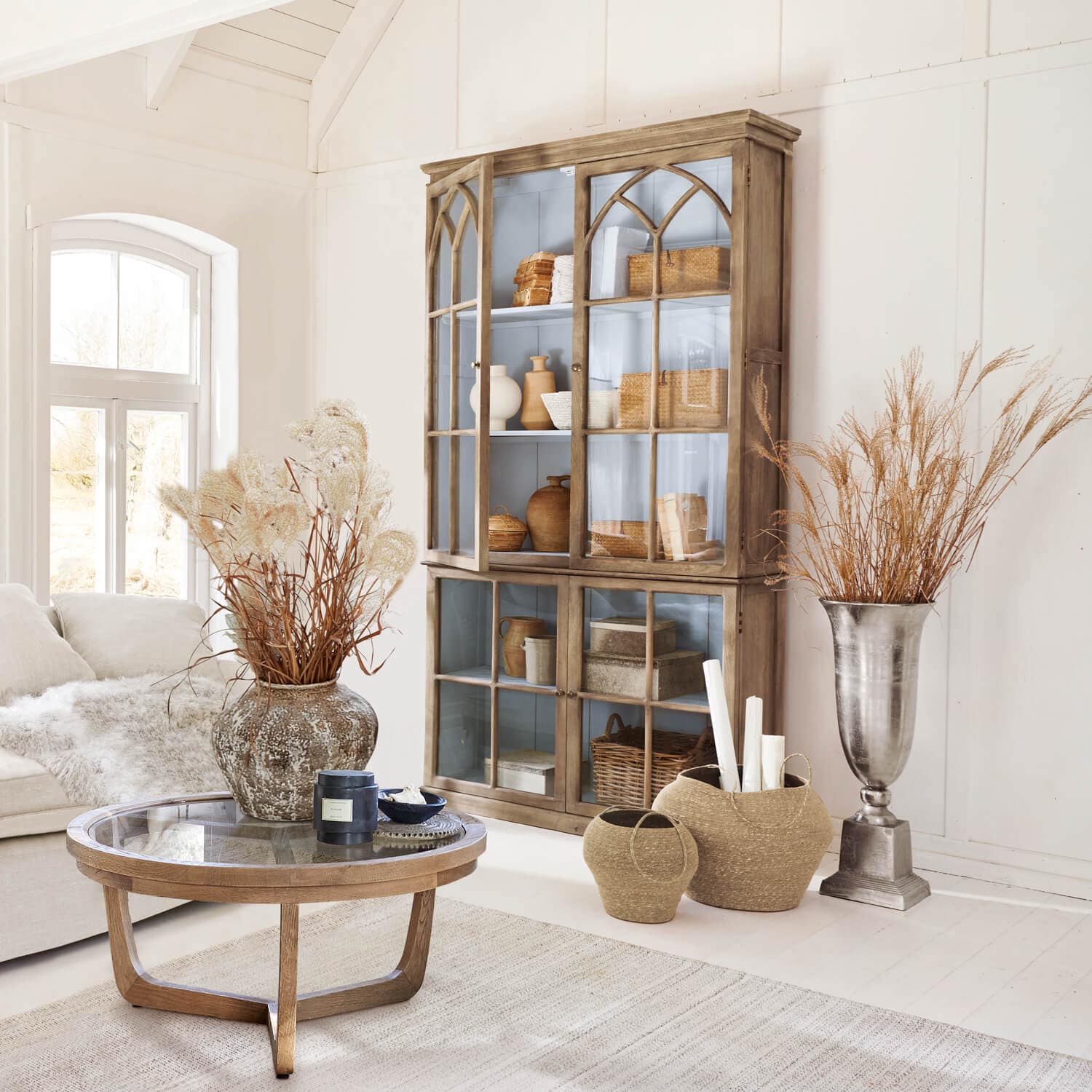 Gemütliches Wohnzimmer mit Holzvitrine, Glastisch und Dekorationen aus Naturmaterialien in hellen Farben