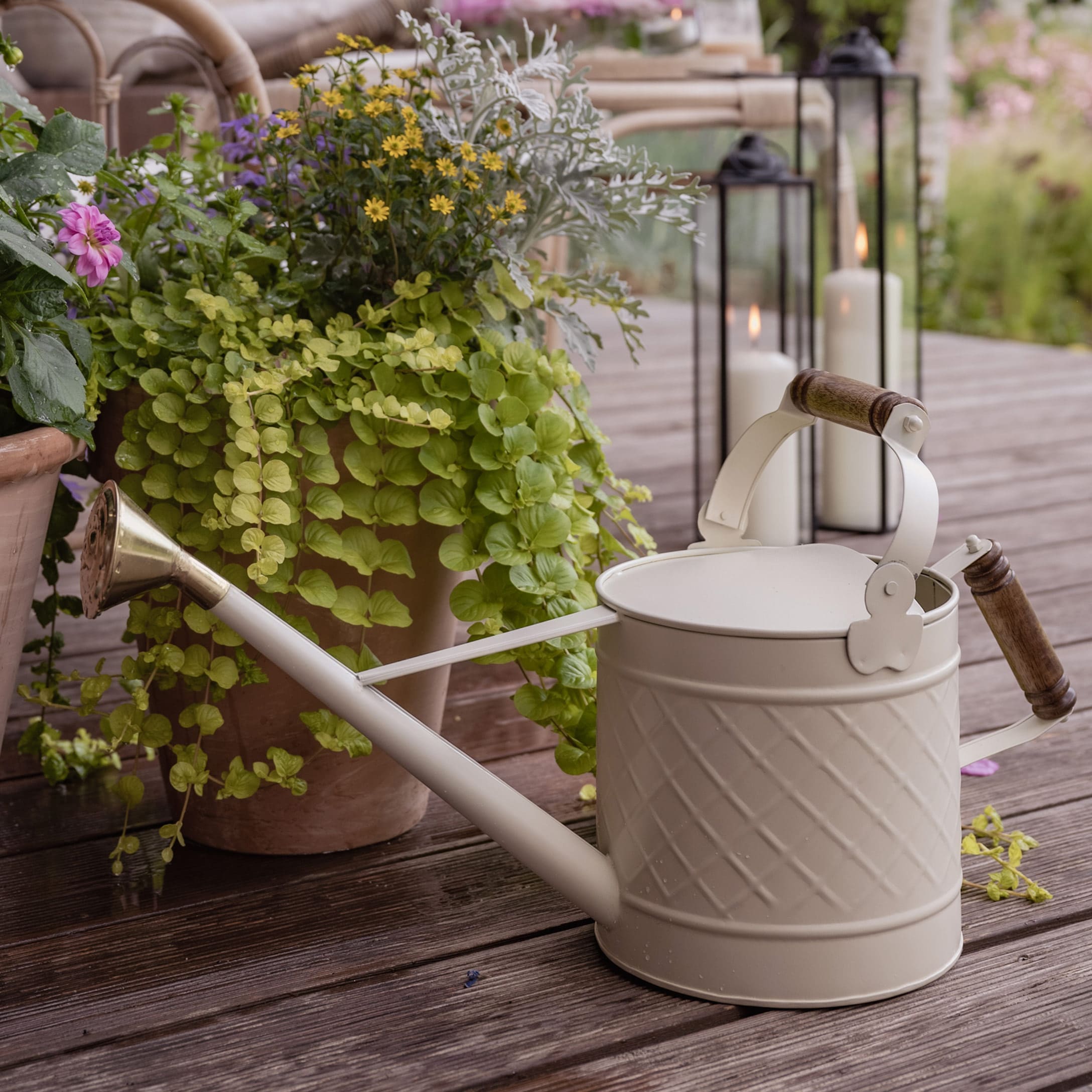 Beige Metall-Giesskanne mit Holzgriff auf Holzterrasse neben Pflanzen und Kerzen
