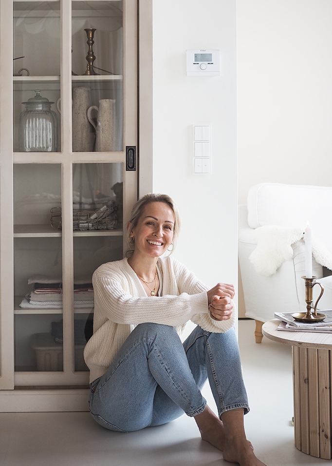 Lächelnde Frau in weissem Pullover und Jeans sitzt auf dem Boden in einem hellen, gemütlichen Raum.