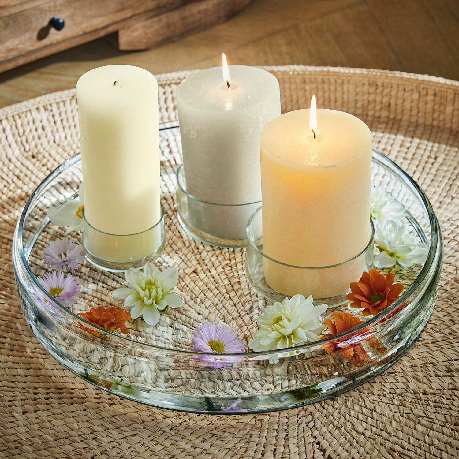 Kerzenteller Marton Glasplatte mit drei pastellfarbenen Kerzen und Blumen auf geflochtenem Tisch