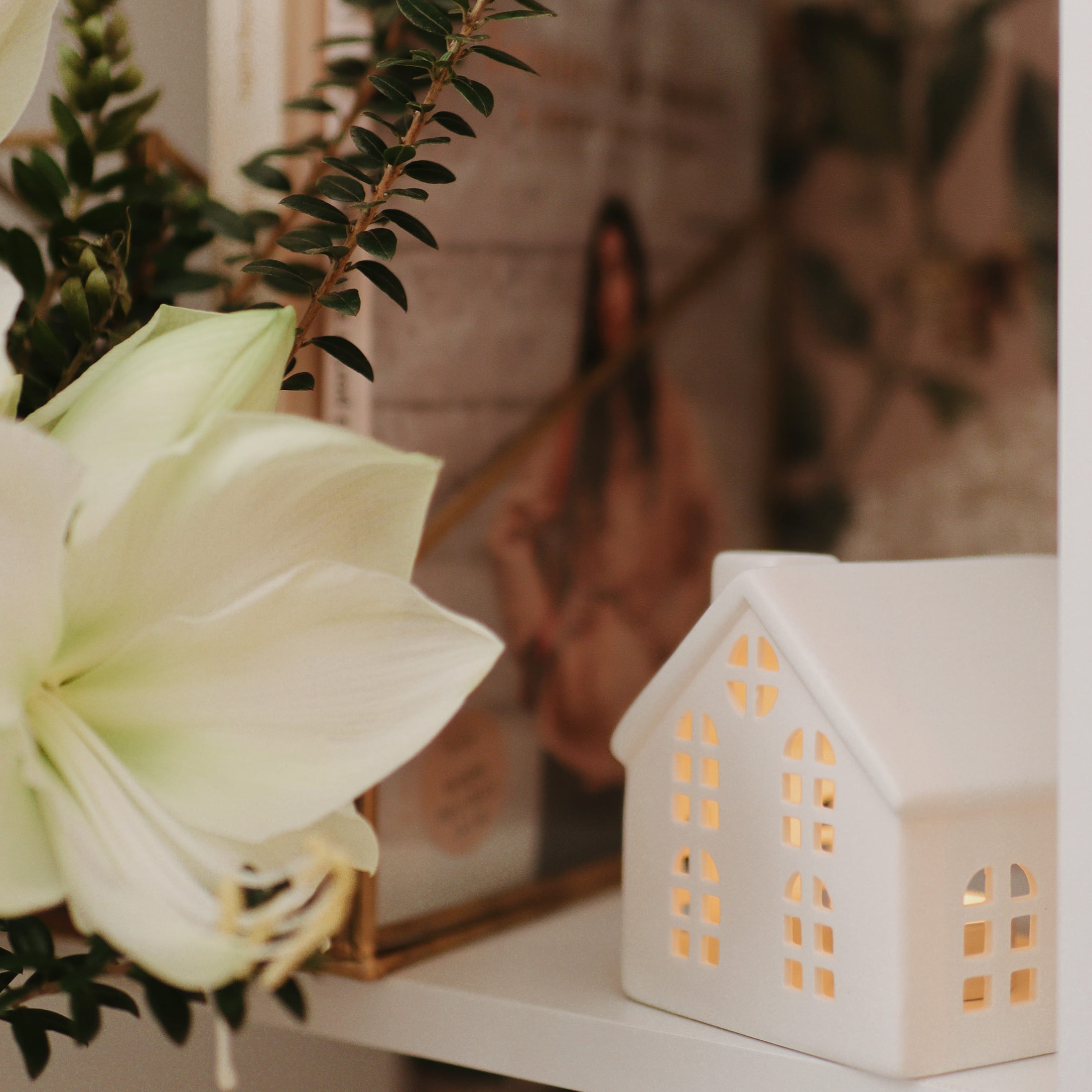 Weisse Keramikhaus-Dekoration mit Beleuchtung und weissen Blumen auf dem Regal, festliche Stimmung