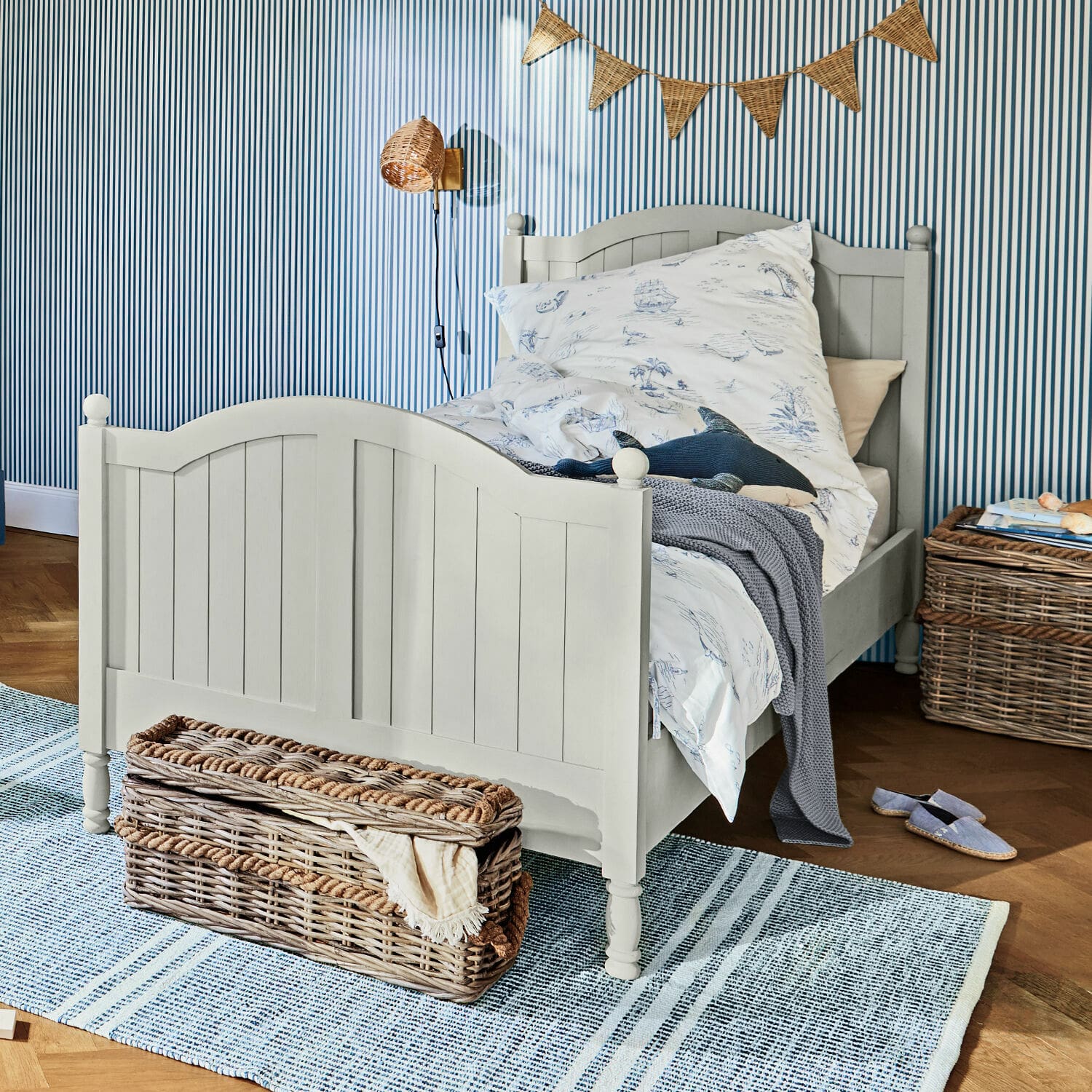 Bett Alarion Graues Kinderbett aus Holz mit maritim gemusterter Bettwäsche und Korb aus Weide auf Teppichboden