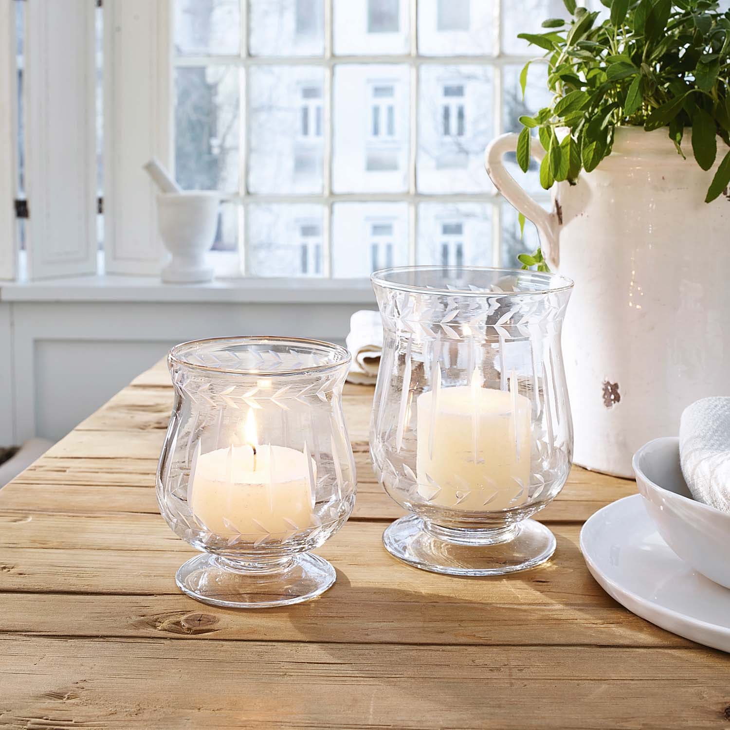 Zwei Glaswindlichter mit weissen Kerzen auf Holztisch in hellem Raum