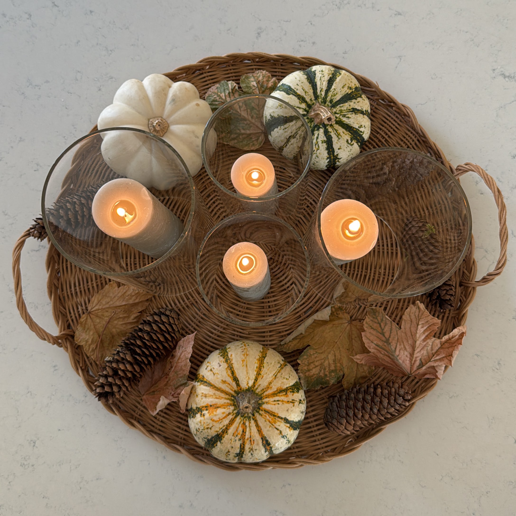Herbstliches Ensemble mit weissen Kürbissen, Tannenzapfen und brennenden Kerzen in Glas auf einem geflochtenen Tablett