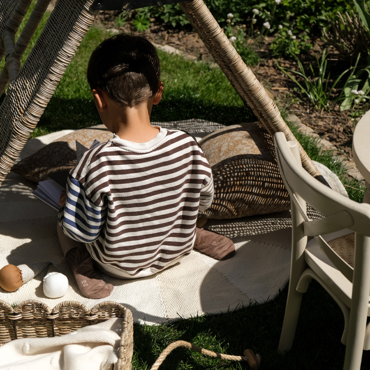 Kind im gestreiften Pullover sitzt auf Teppich unter Weidenzelt im Garten, entspannte Spielsituation