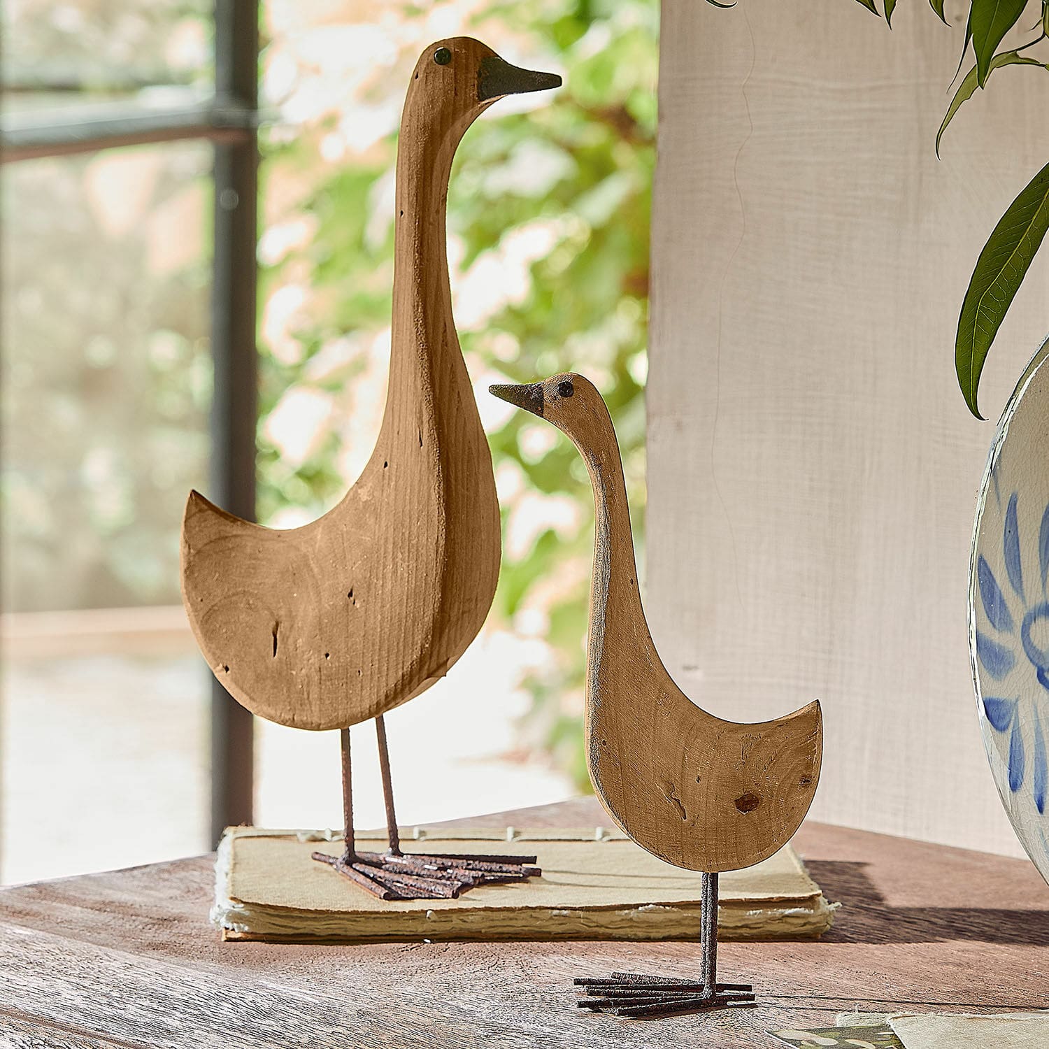 Deko-Gans 2er Set Amiraz Holzdekorationen in Vogelgestalt in Naturfarbe auf Metallfüssen auf einem Tisch platziert.