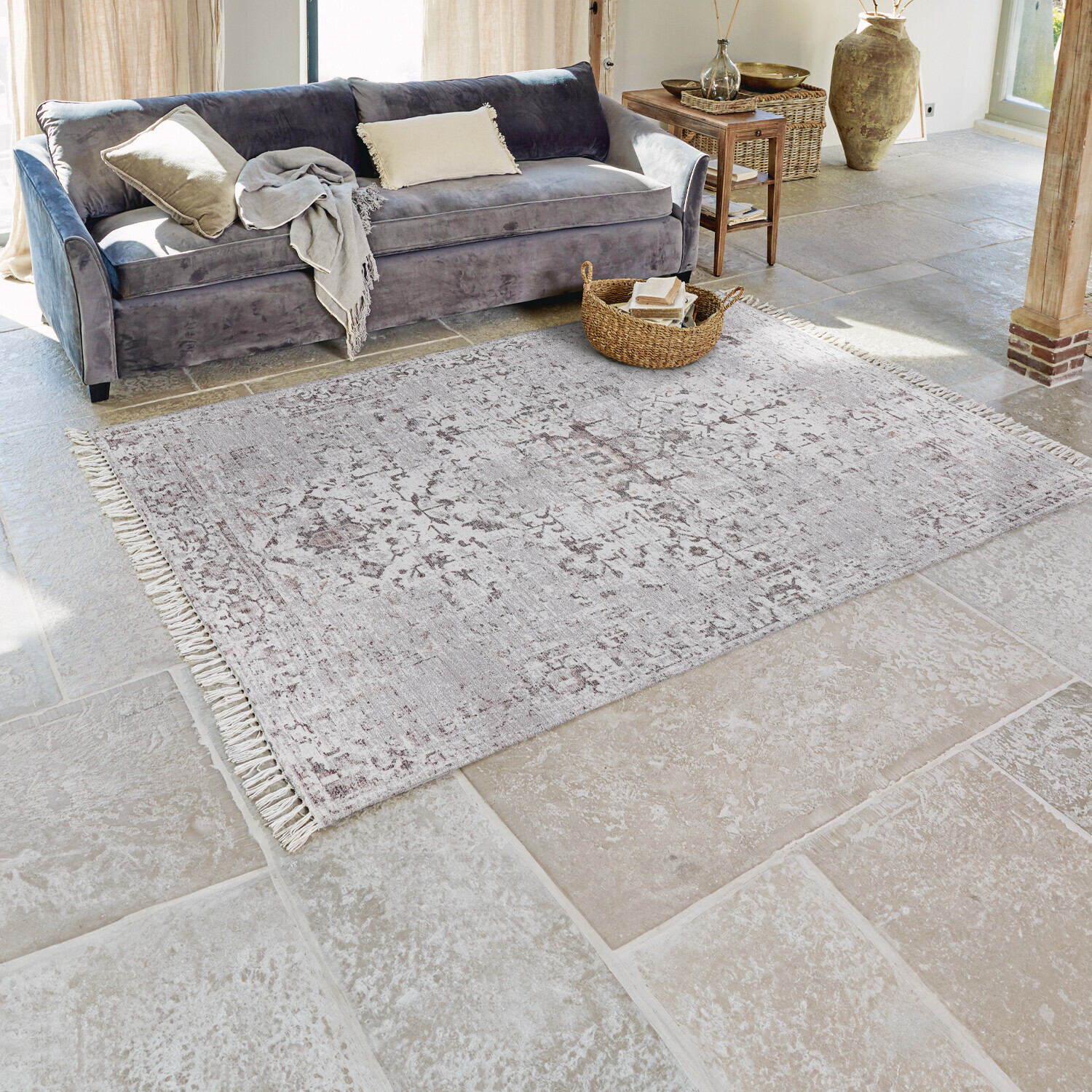 Grauer Teppich mit feinem Muster und Fransen auf Steinboden in hellem Wohnzimmer mit grauem Sofa