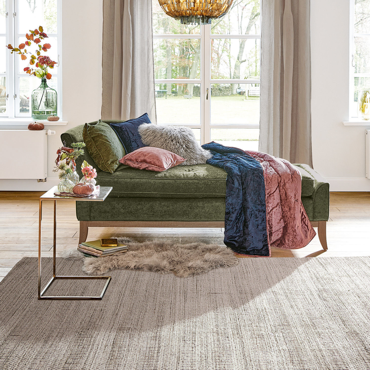 Chaiselongue Alphonse Grüne Samt-Chaiselongue mit Kissen und rosa-blauer Decke in hellem Wohnzimmer mit grossem Fenster