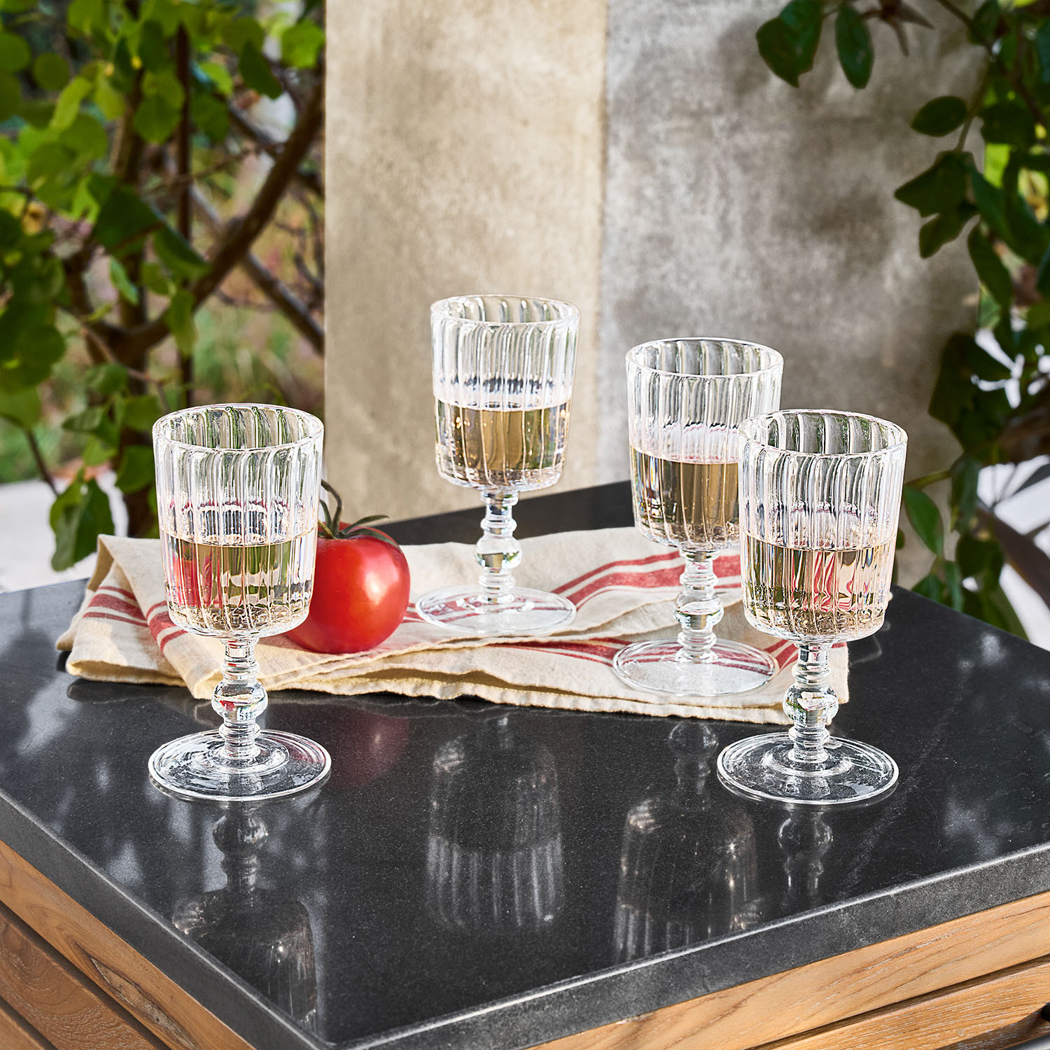 Vier gerillte Gläser aus klarem Glas mit Weisswein auf schwarzer Arbeitsplatte neben einer Tomate