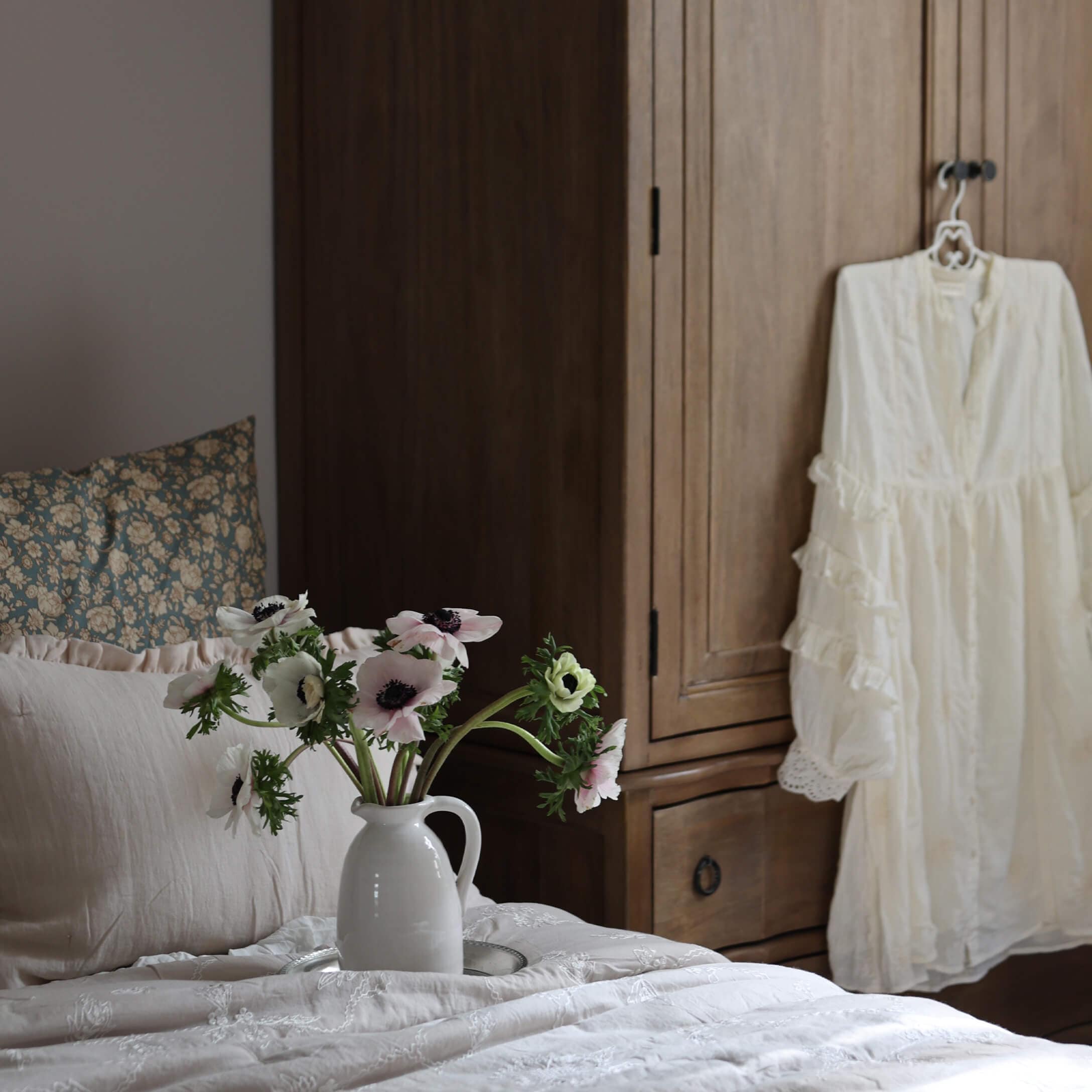 Weisser Keramikkrug mit Blumen auf hellem Bett vor Holzschrank im Schlafzimmer