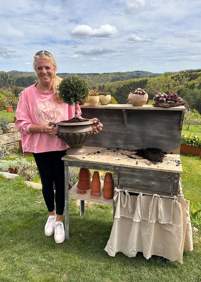 Frau im rosa Pullover hält einen Topf mit grünem Strauch an einem alten Gartentisch vor grünem Landschaftshintergrund.