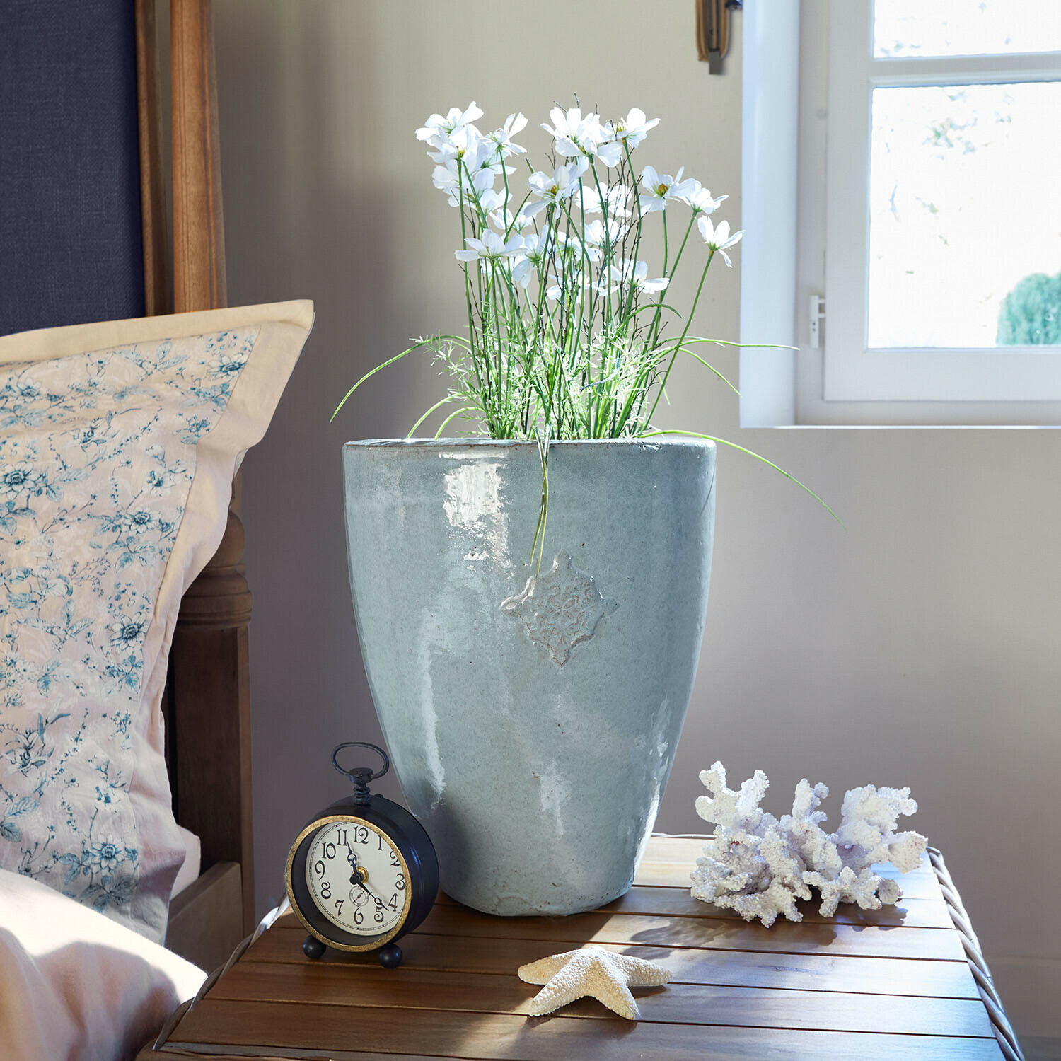 Übertopf Serenzia Grauer Keramikblumentopf mit weissen Blumen auf Holztisch neben Wecker und Meeresdekorationen