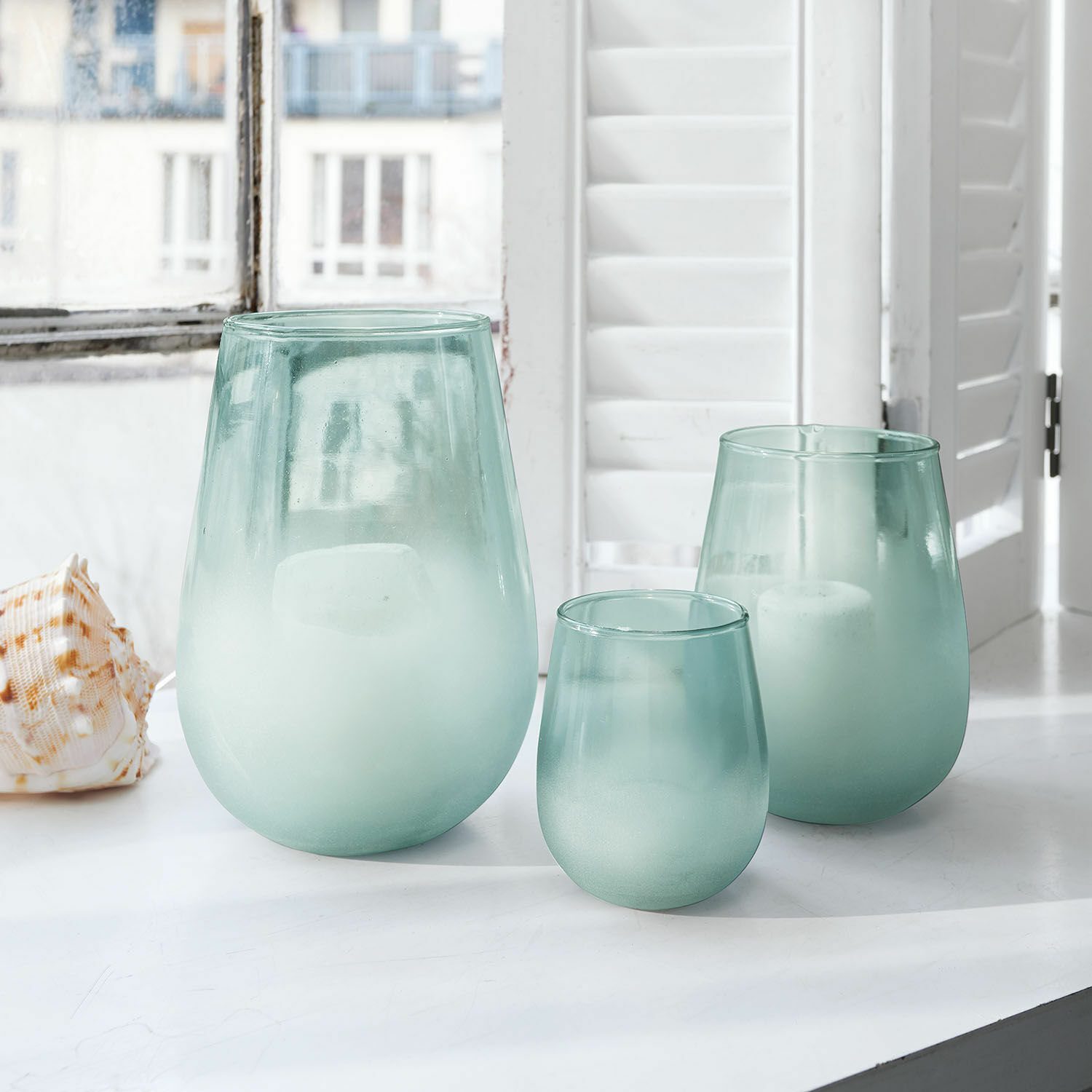 Windlicht 3er Set Miristelle Drei matte, grünliche Glaswindlichter auf dem weissen Fensterbrett mit Kerzen im Innern