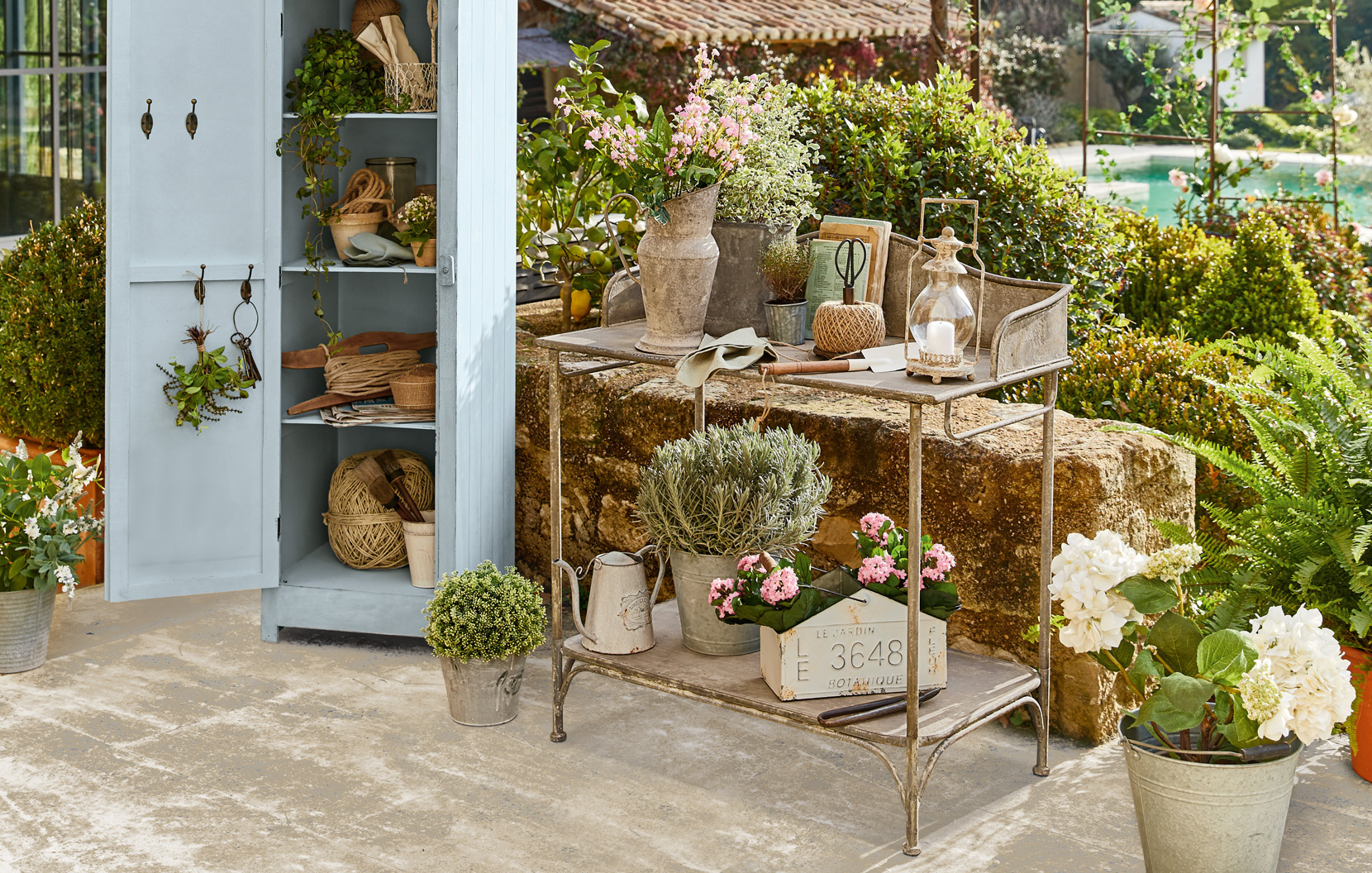 Metall-Gartenwagen mit Shabby-Chic-Dekorationen und Blumen auf der Terrasse neben einem blauen Schrank.