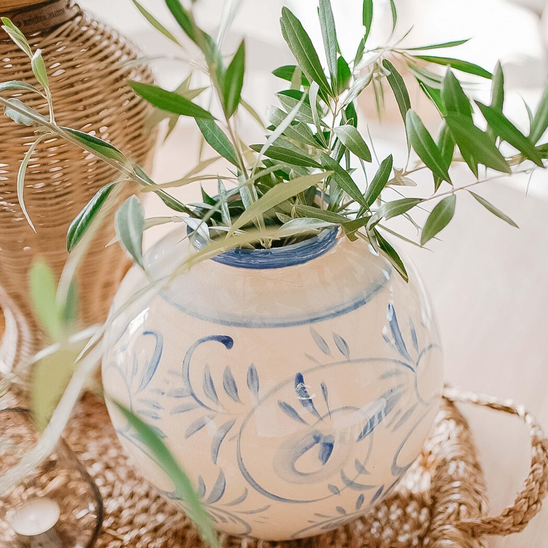 Weisse Keramikvase mit blauem Muster und grünen Blättern auf einem geflochtenen Tablett, Frühlingsdekoration