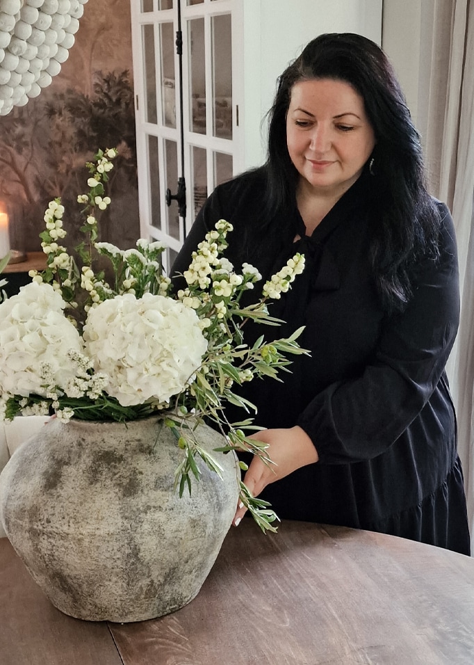 Frau arrangiert weissen Blumenstrauss in grosser grauer Keramikvase auf Holztisch.