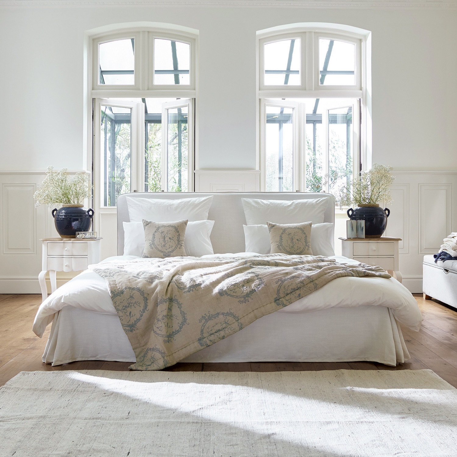 Bett Aubrianne Gemütliches Bett mit hellem Bezug und beigefarbener Decke mit Muster in hellem Schlafzimmer mit grossen Fenstern