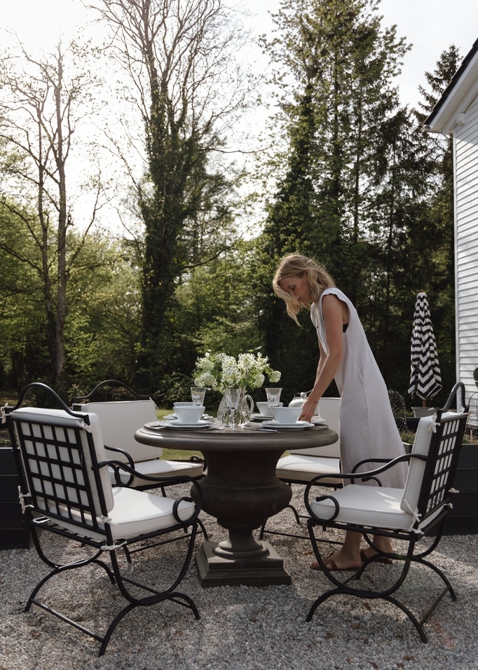 Garten mit rundem Steintisch und schwarzen Metallstühlen mit weissen Polstern, Frau im weissen Kleid.