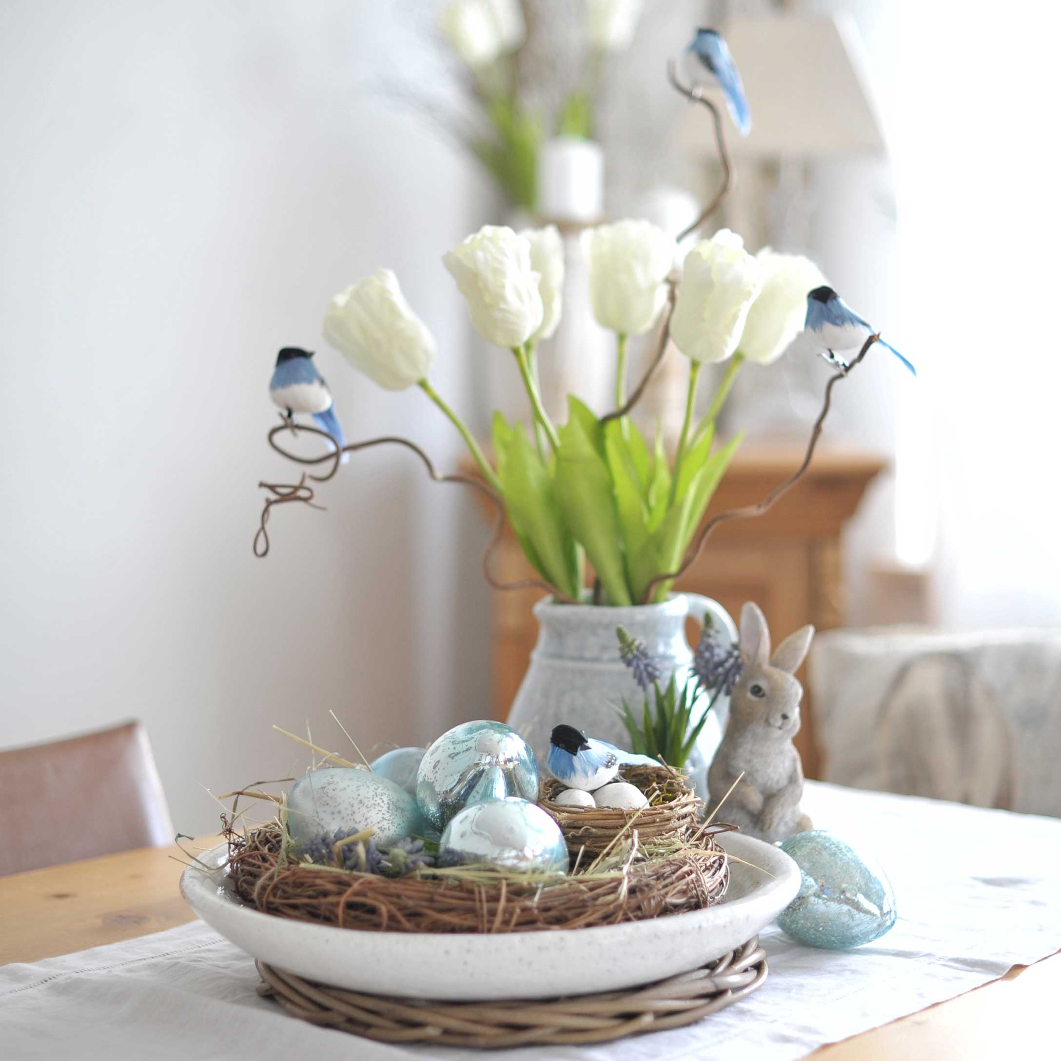 Dekorativer Kranz mit blauen Eiern und Vögeln auf Tisch mit weissen Tulpen und Hasenfigur