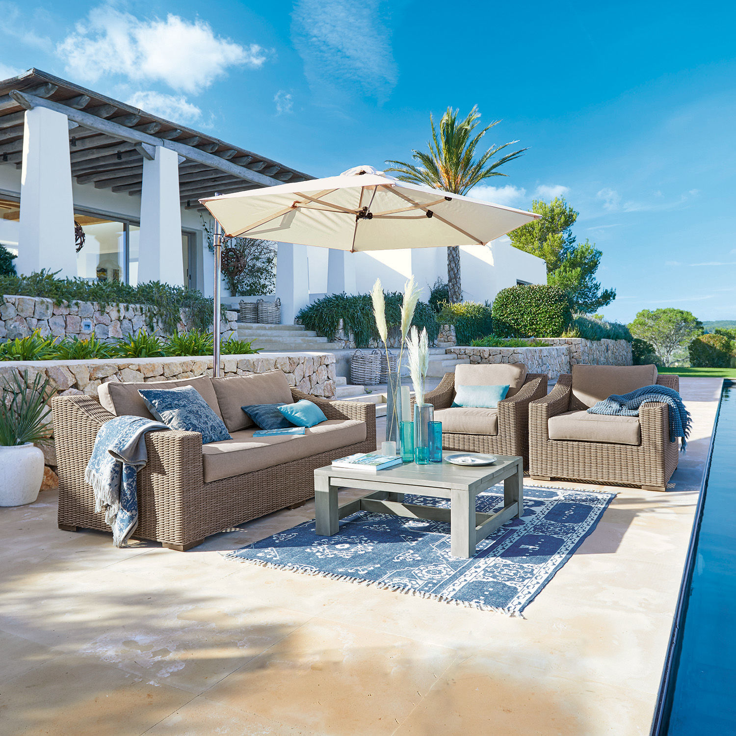 Sitzgruppe Chestingdale Beiges Rattan-Gartenmöbel-Set mit Sonnenschirm und blauem Teppich auf sonniger Terrasse
