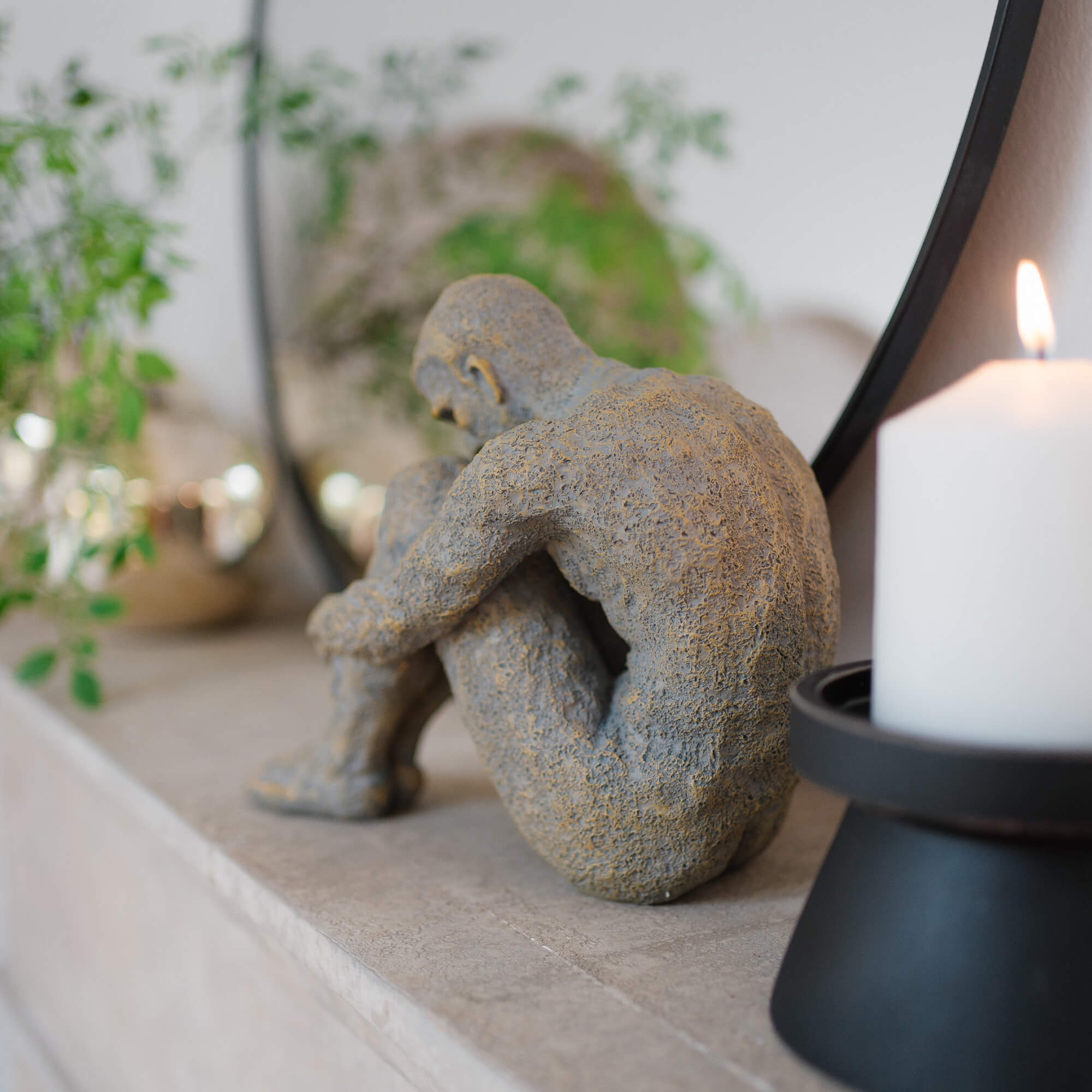 Graue Steinskulptur einer sitzenden Figur auf Marmorablage neben weisser Kerze