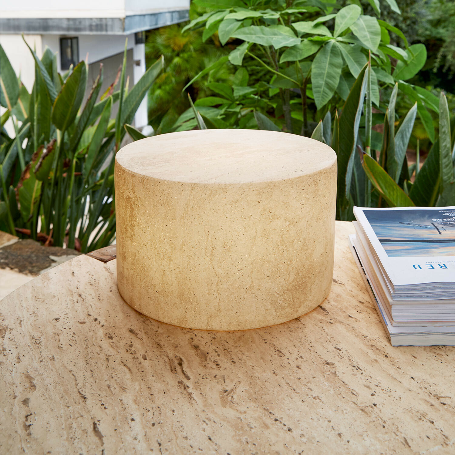 Wiederaufladbare LED-Lampe Marévos Runder Beistelltisch aus beigem Travertin auf Terrasse mit grünen Pflanzen, Naturstein