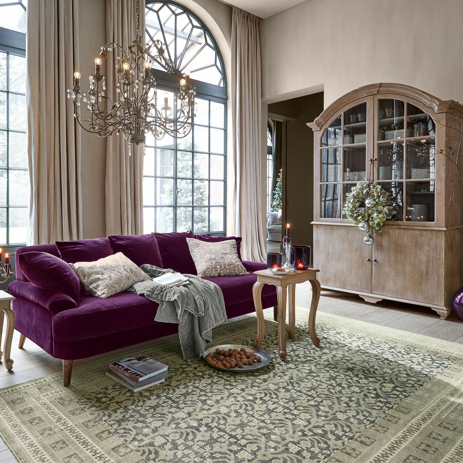 Teppich Nadimah Wohnzimmer mit violettem Samtsofa und Holzvitrine vor grossen Fenstern und gemustertem Teppich