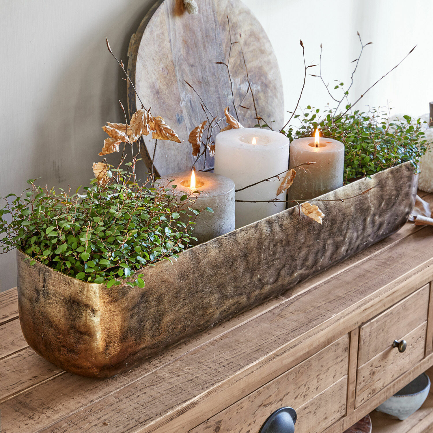 Schale Skye Dekorativer goldener Metall-Blumenkasten mit grünen Pflanzen und drei Kerzen auf Holztisch.