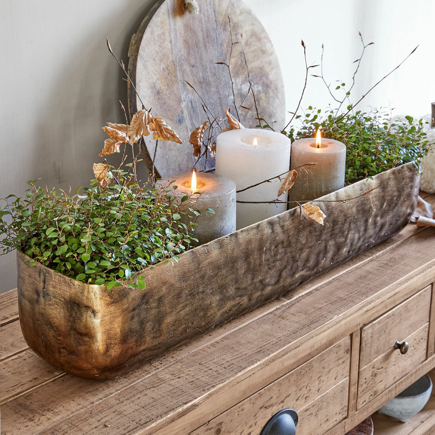 Schale Skye Dekorativer goldener Metall-Blumenkasten mit grünen Pflanzen und drei Kerzen auf Holztisch.