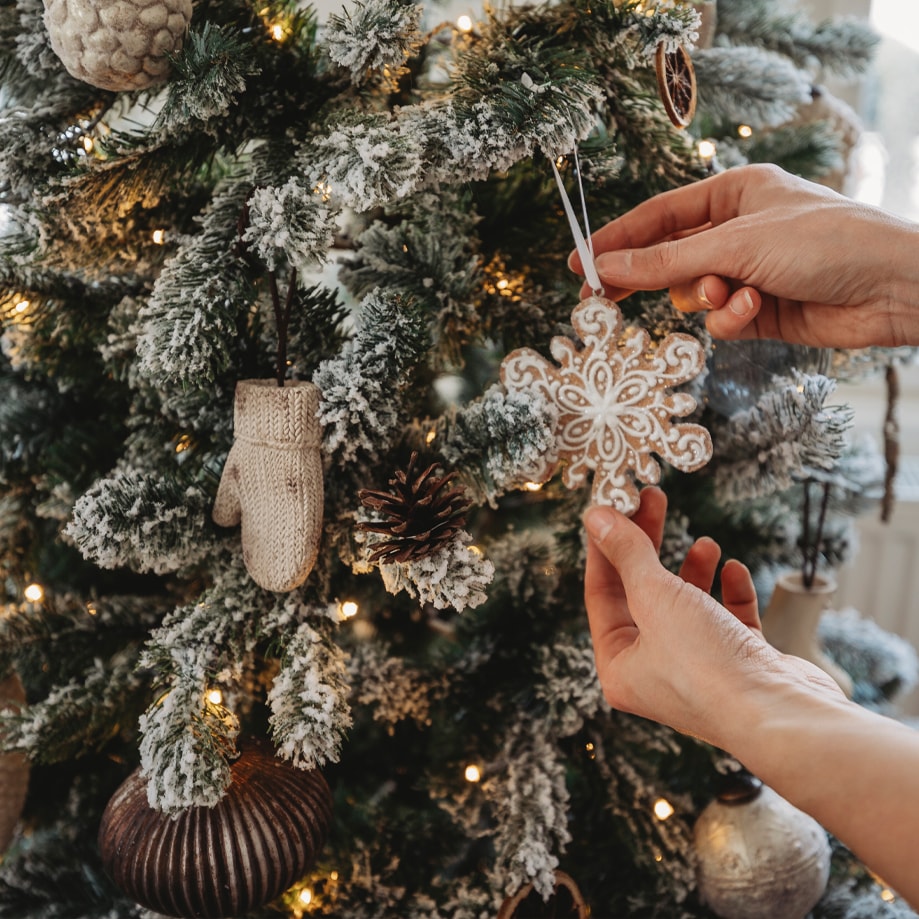 Hände hängen ein Schneeflockenornament an einen schneebedeckten Weihnachtsbaum mit Lichterkette.