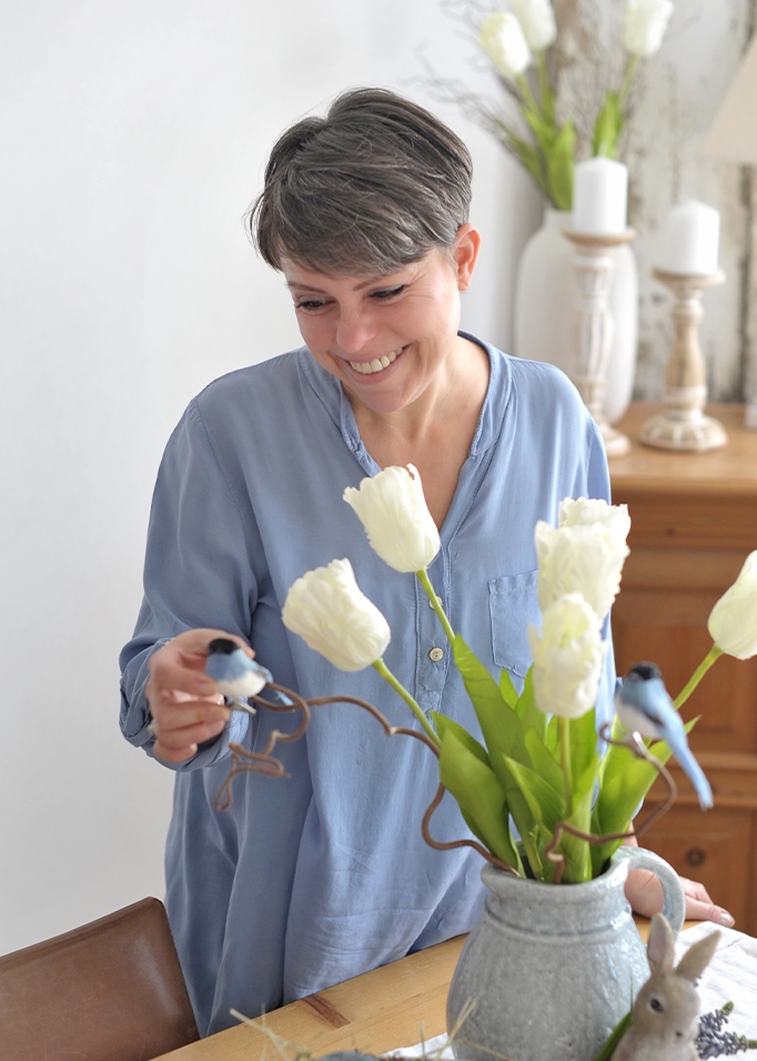Frau im blauen Hemd dekoriert den Tisch mit einer grauen Keramikvase, weissen Tulpen und Deko-Vögeln.