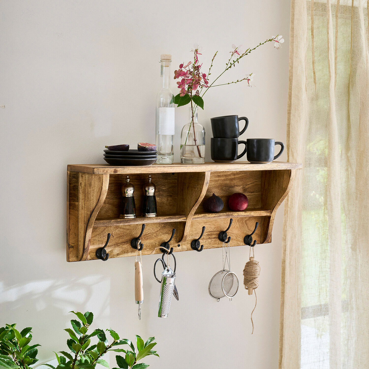 Wandregal aus Holz mit Haken, schwarzen Tassen, Tellern und Blumenarrangement in hellem Raum