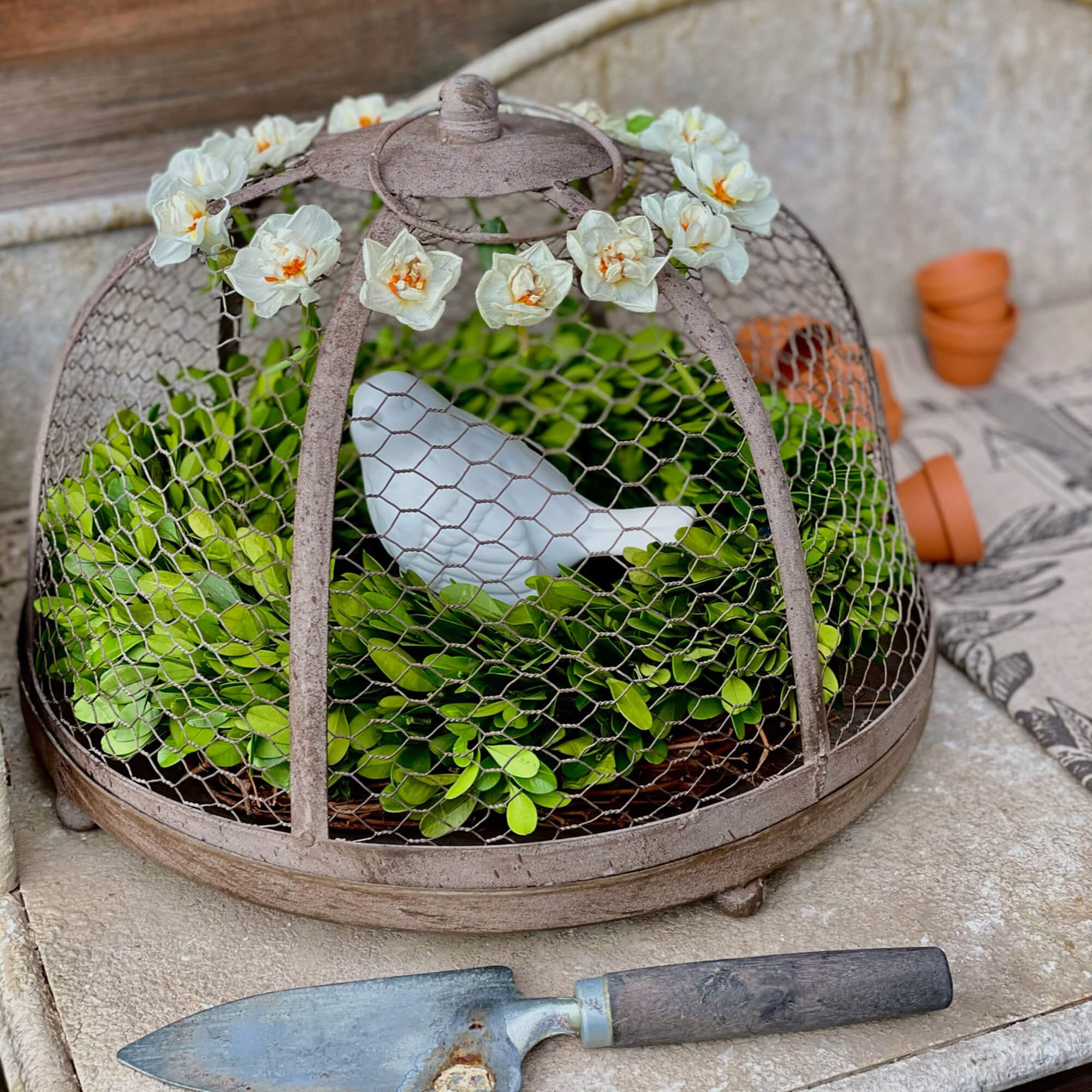 Metall-Gartendekoration mit grünem Laub und weissem Keramikvogel unter blumengeschmücktem Käfig