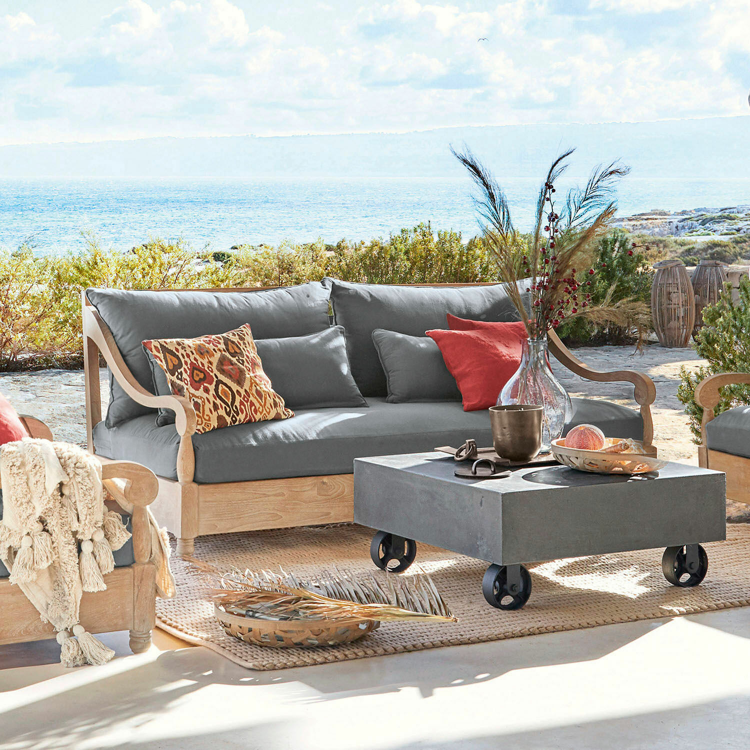 Sofa Montcassin Outdoor-Sofa aus hellem Holz mit grauen Polstern und bunten Dekokissen auf Terrasse mit Meerblick