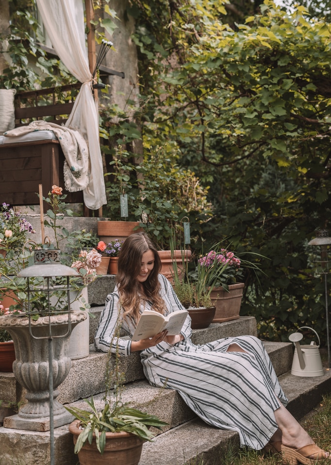 Frau im gestreiften Kleid liest Buch auf Steintreppe, umgeben von Blumentöpfen im Garten.