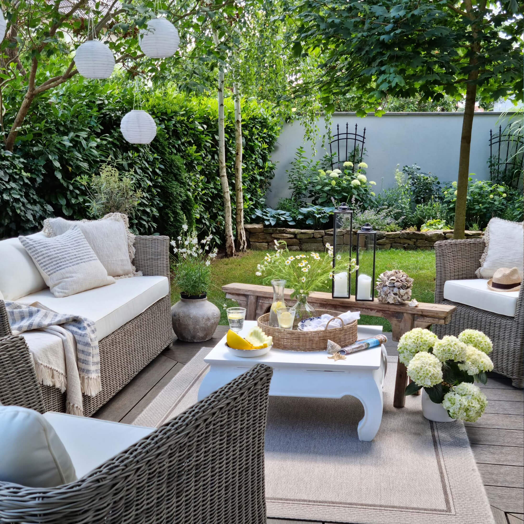 Gemütliche Gartenecke mit grauen Rattanmöbeln und weissen Kissen, Dekorationen und grünen Pflanzen.